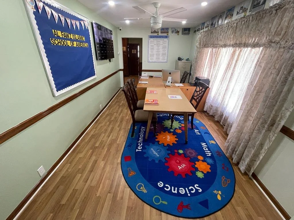 A meeting or classroom room with a table, multiple chairs, curtains, a bulletin board, and a rug with colorful gears and educational words. The room appears to be part of Jalal Fawz Islamic School of America.