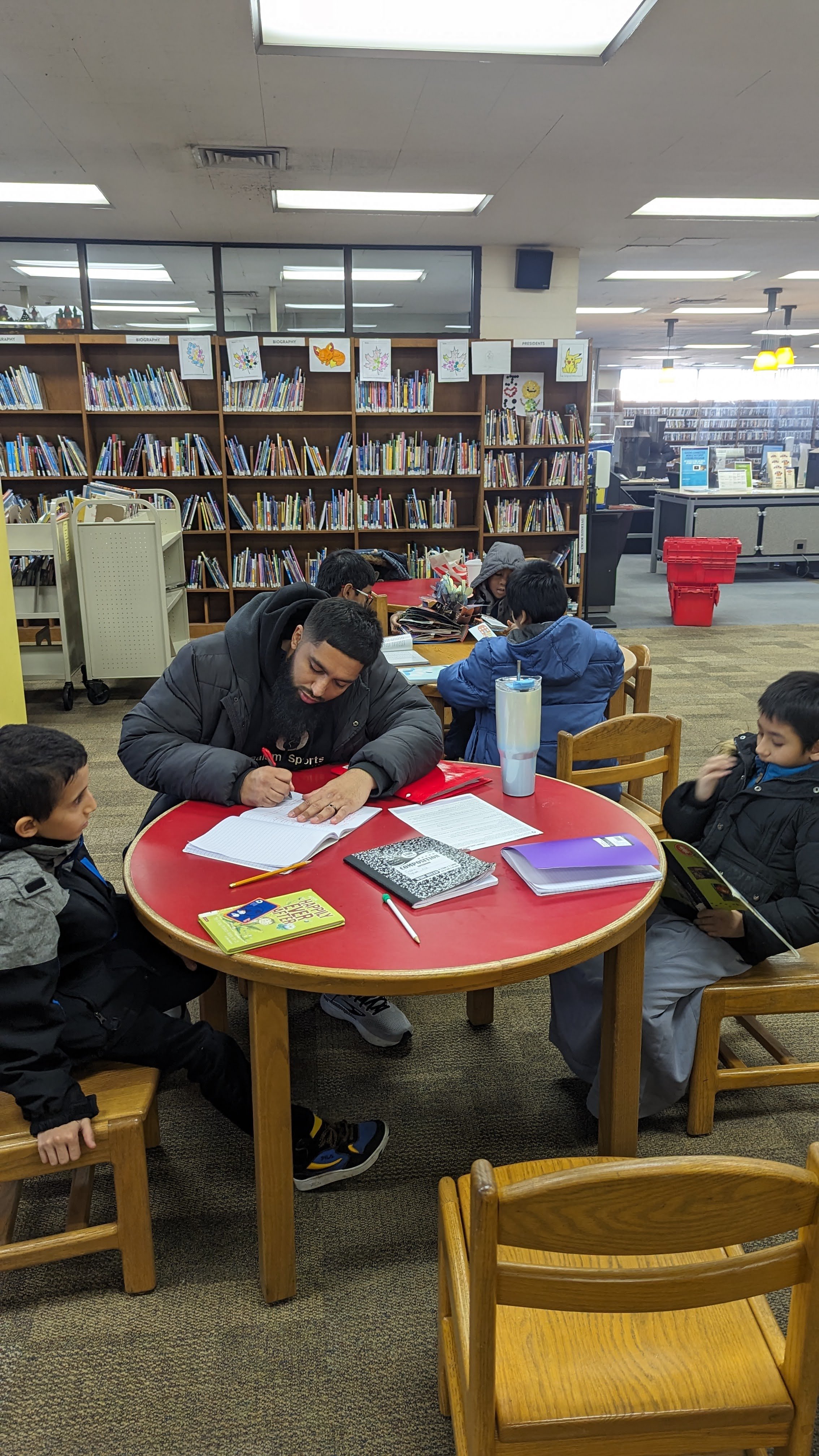 A man and two children sitting at a round table in a library, with books and papers in front of them, and other children studying in the background.