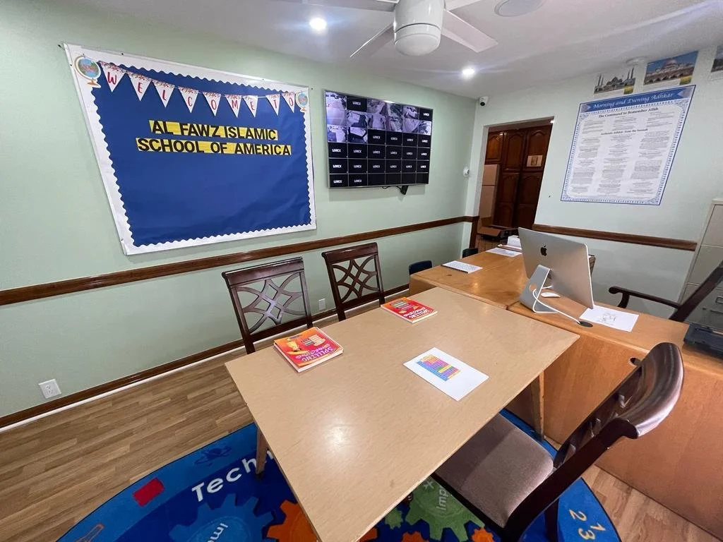 Conference room with a blue bulletin board that says 'WELCOME' and 'ALL FAWZIS AIC SCHOOL OF AMERICA', a wall-mounted security monitor, a wooden desk with a computer and books, and globe-themed decor.