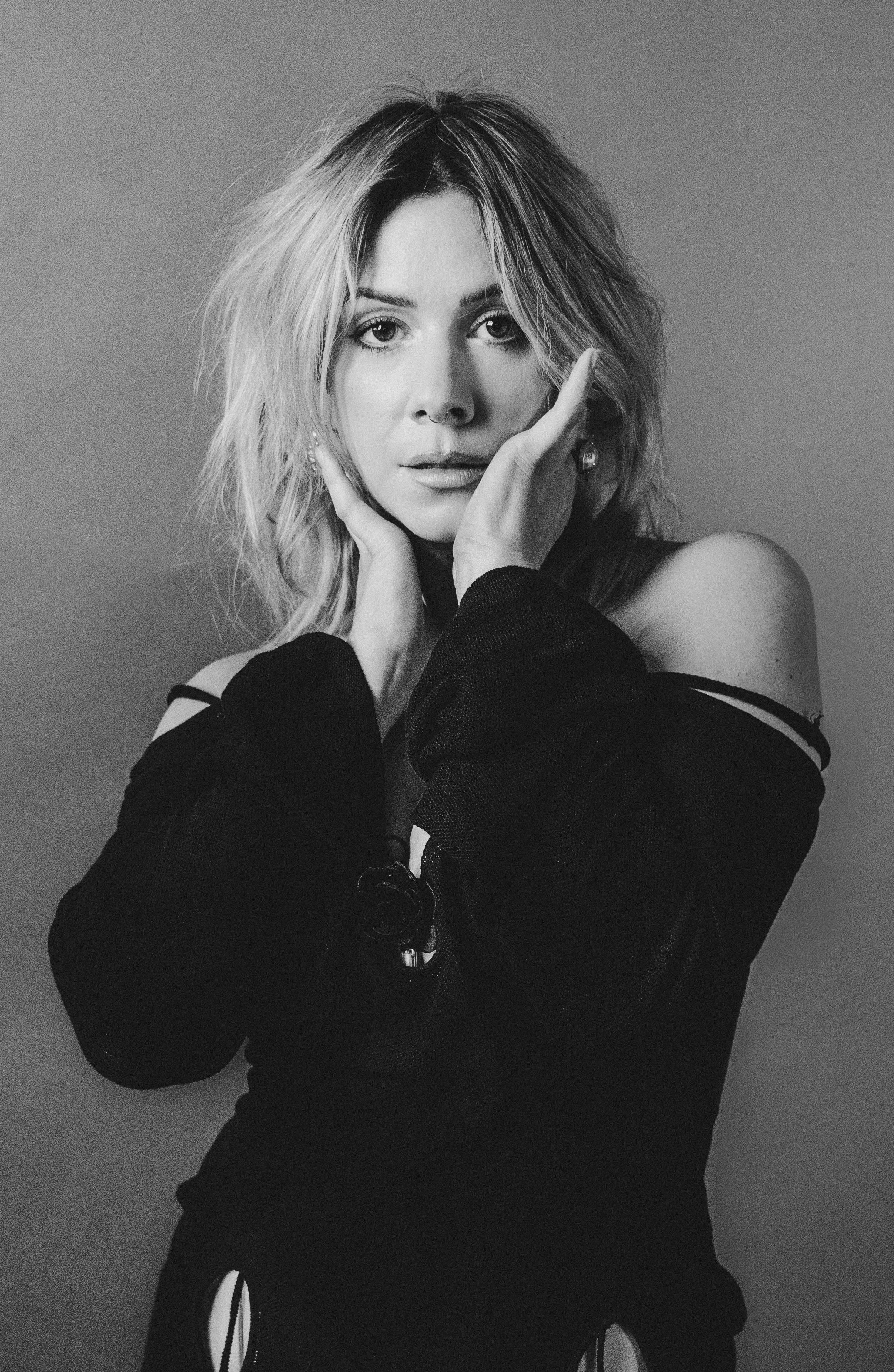 Black and white portrait of a woman with wavy hair, touching her face with her hands, wearing a dark, off-the-shoulder top.