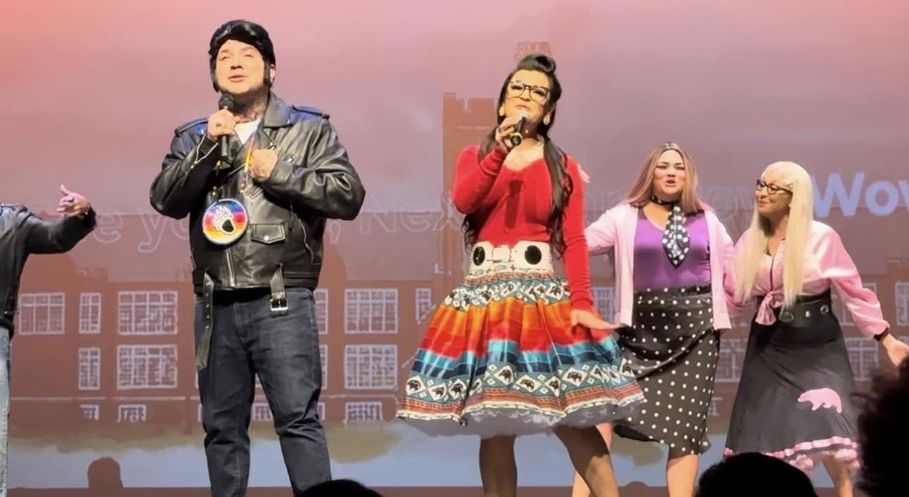 A group of four performers on stage, holding microphones, with a pinkish background and a faint cityscape image.