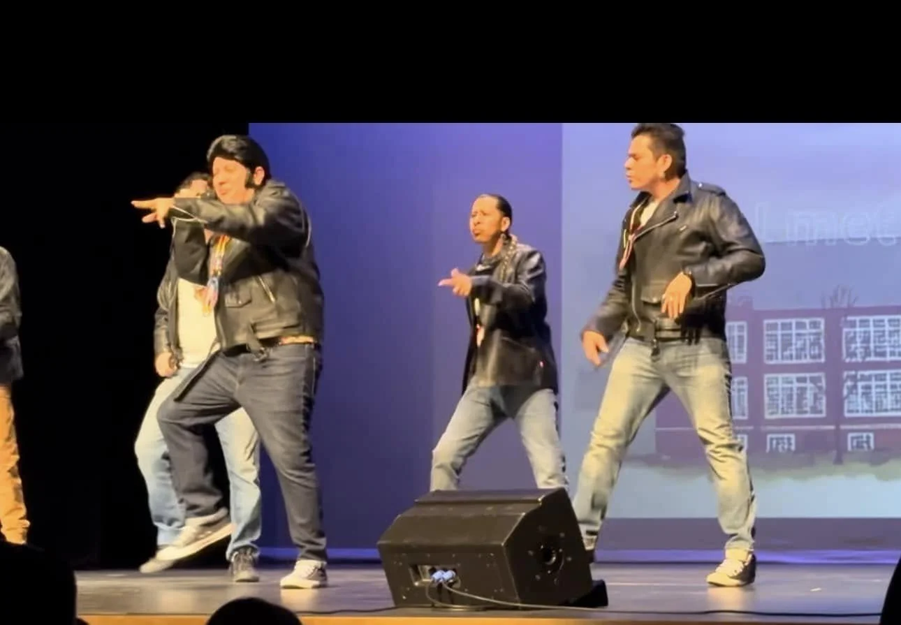 Group of men performing a dance on stage, wearing black leather jackets and jeans, with a backdrop of a school building.