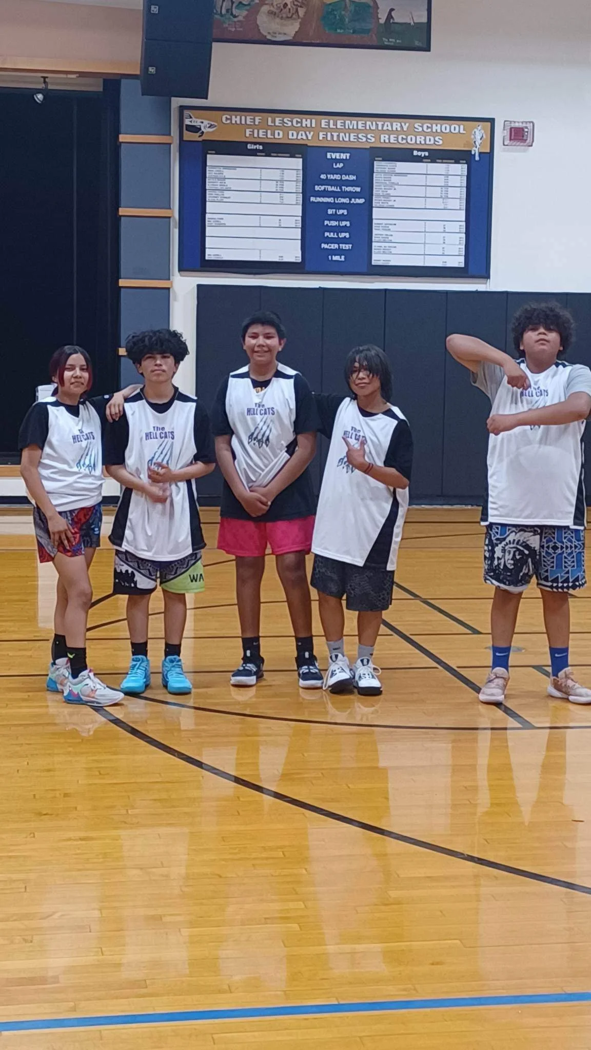 Five children standing on a basketball court in front of a scoreboard at Chief Leschi Elementary School. They are wearing sports uniforms for a field day event.
