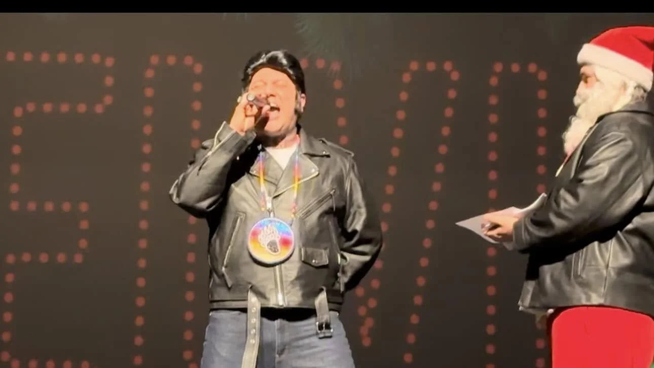 Two men on stage, one in a leather jacket with a Christmas medal, singing into a microphone, and the other in a leather jacket wearing a Santa hat, holding a paper.