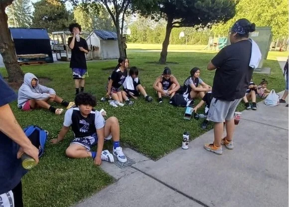 Group of young students in school uniforms sitting on the grass and sidewalk outdoors, with a person standing in front of them, some students holding water bottles.