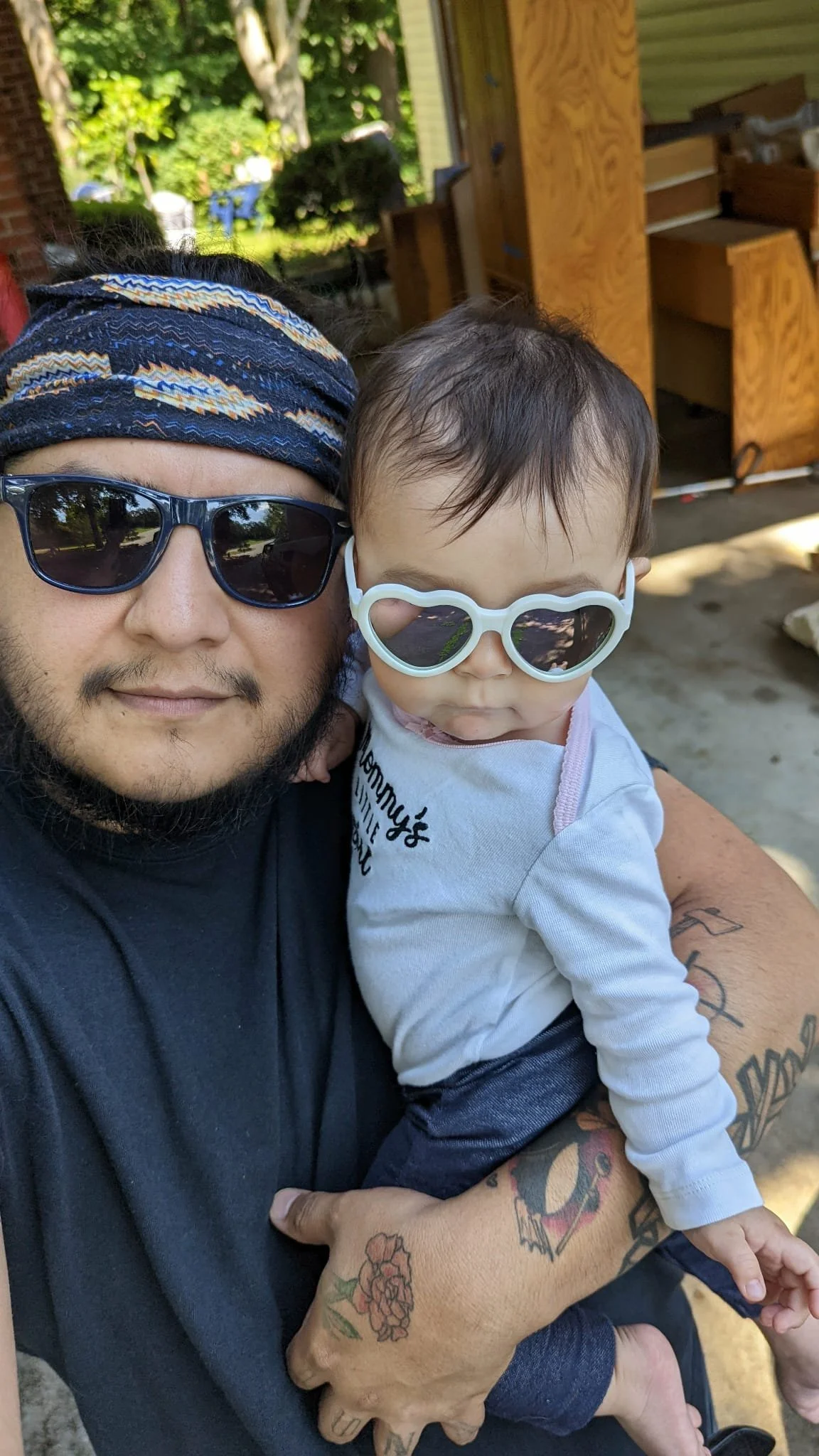 A man wearing black sunglasses and a colorful headband holding a young child with heart-shaped sunglasses outside in a backyard with greenery and a wooden structure.