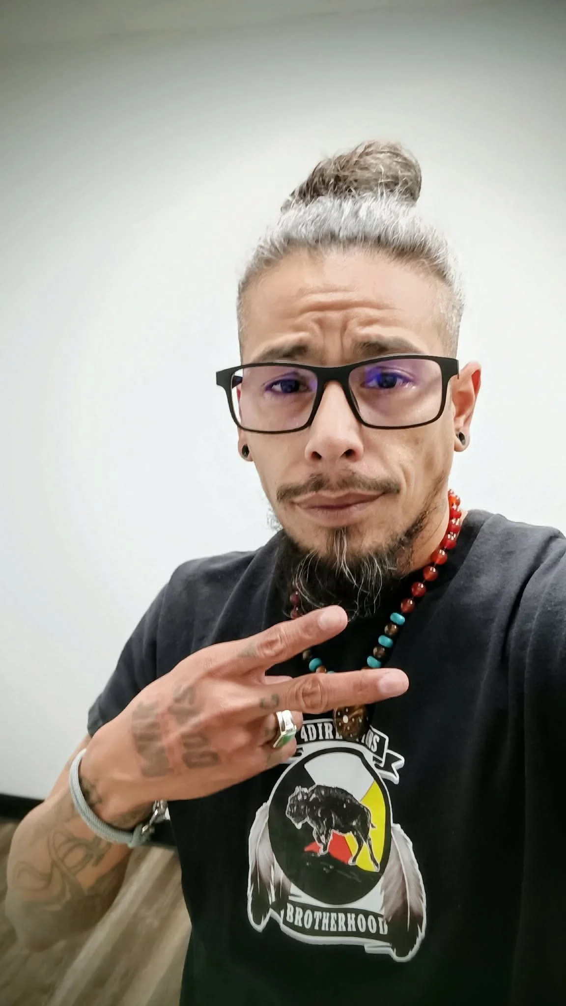 Two smiling women taking a selfie outdoors among green leafy trees. One woman has glasses, dark hair, and a beard, wearing a black T-shirt with a graphic and a beaded necklace. The other woman has glasses, red hair, tattoos on her arms, and is wearing a gray tank top and a cap. She is holding a phone.