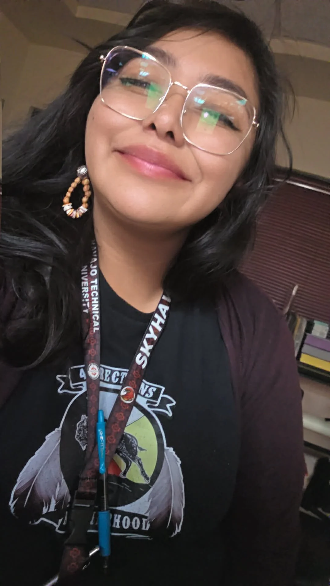 Person taking a selfie in an elevator wearing glasses, a face mask, a black jacket, a badge, and a colorful traditional necklace and earrings.
