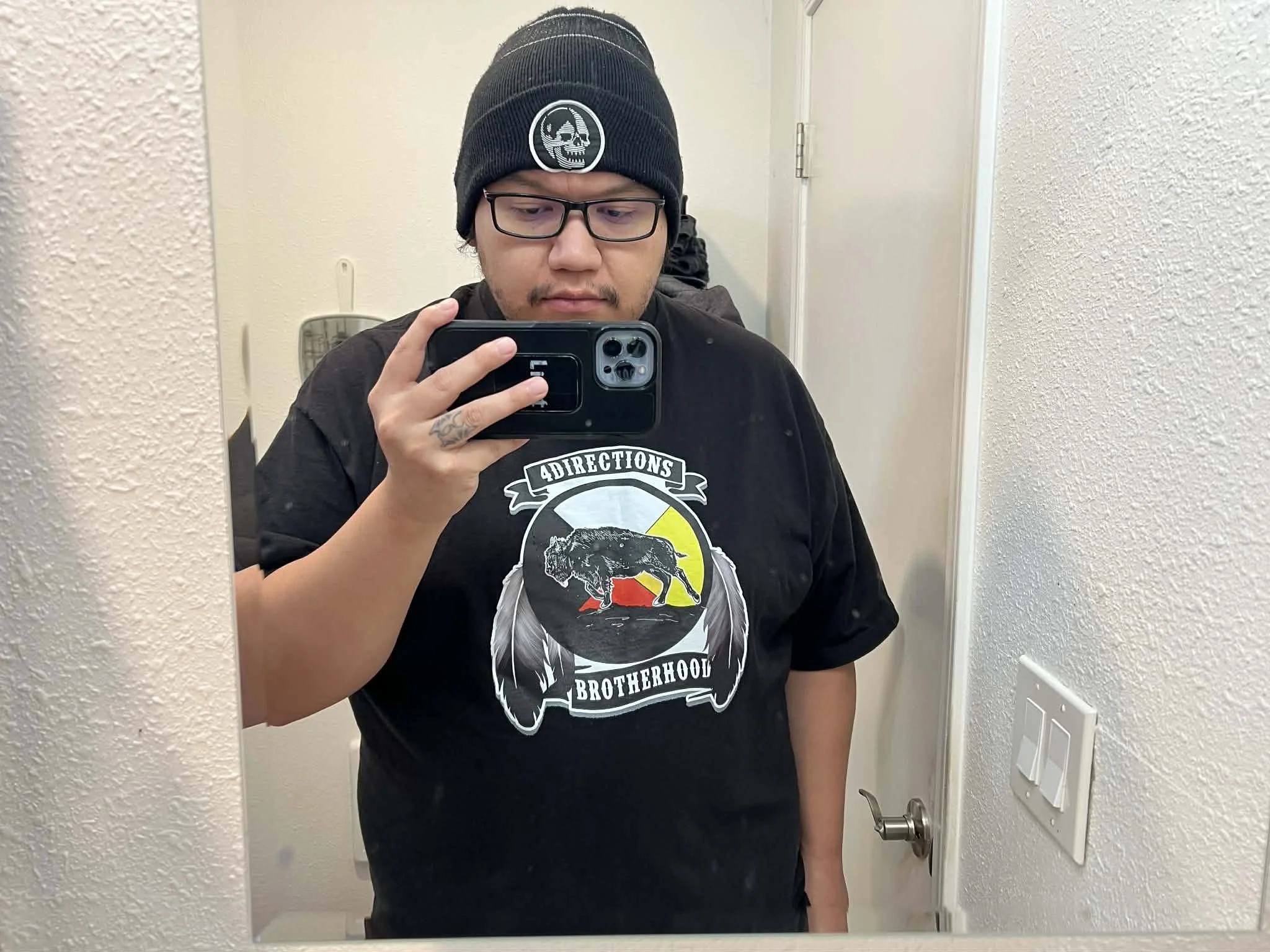 A person with long braided hair, glasses, and wearing a black t-shirt taking a mirror selfie in a bathroom with a smartphone.