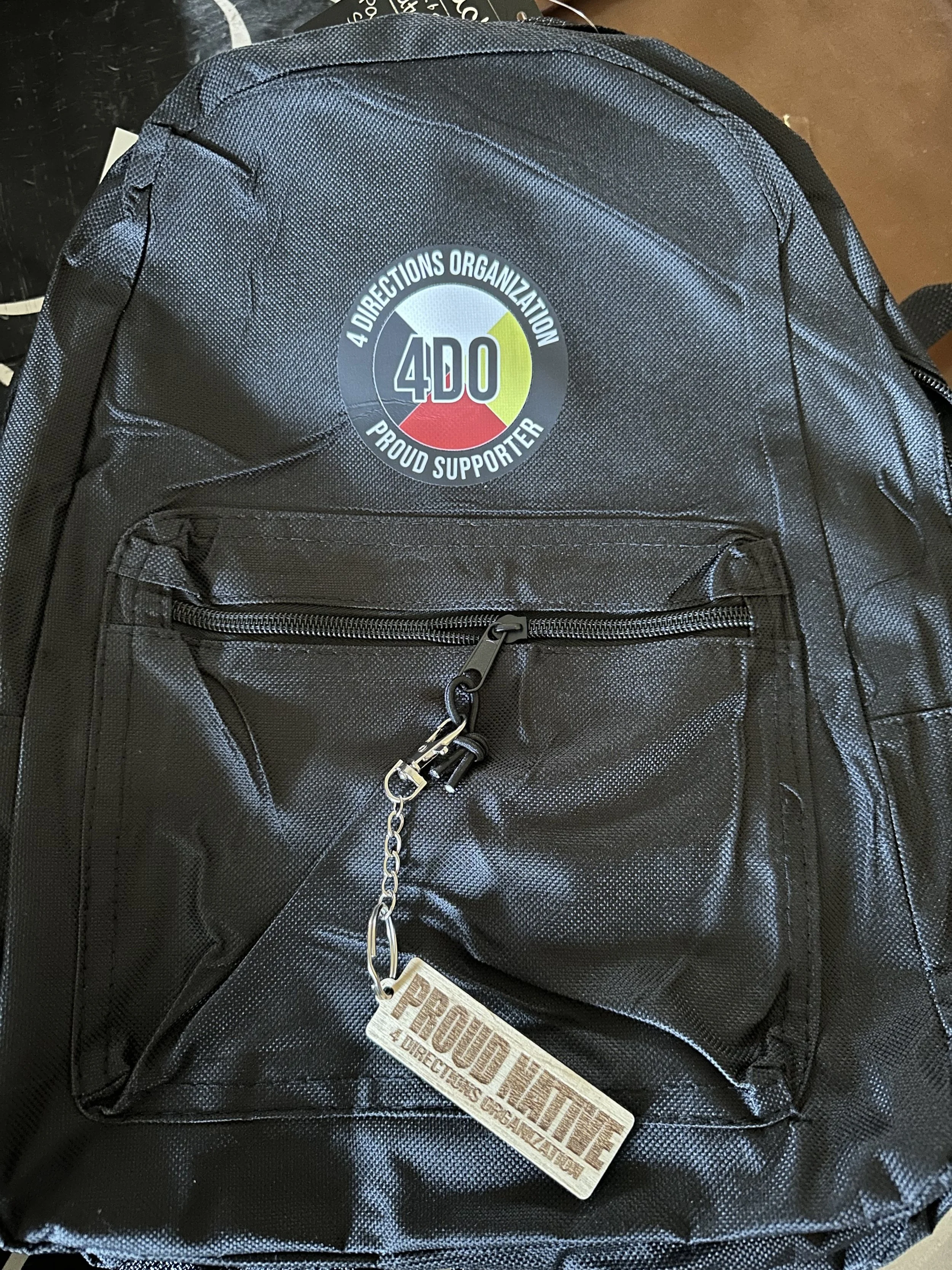 Black backpack with a round sticker featuring the logo of 4D0 and the words 'A Directions Organization Proud Supporter'. A metal keychain attached to a zipper reads 'Proud Family - 4 Directions Organization'.