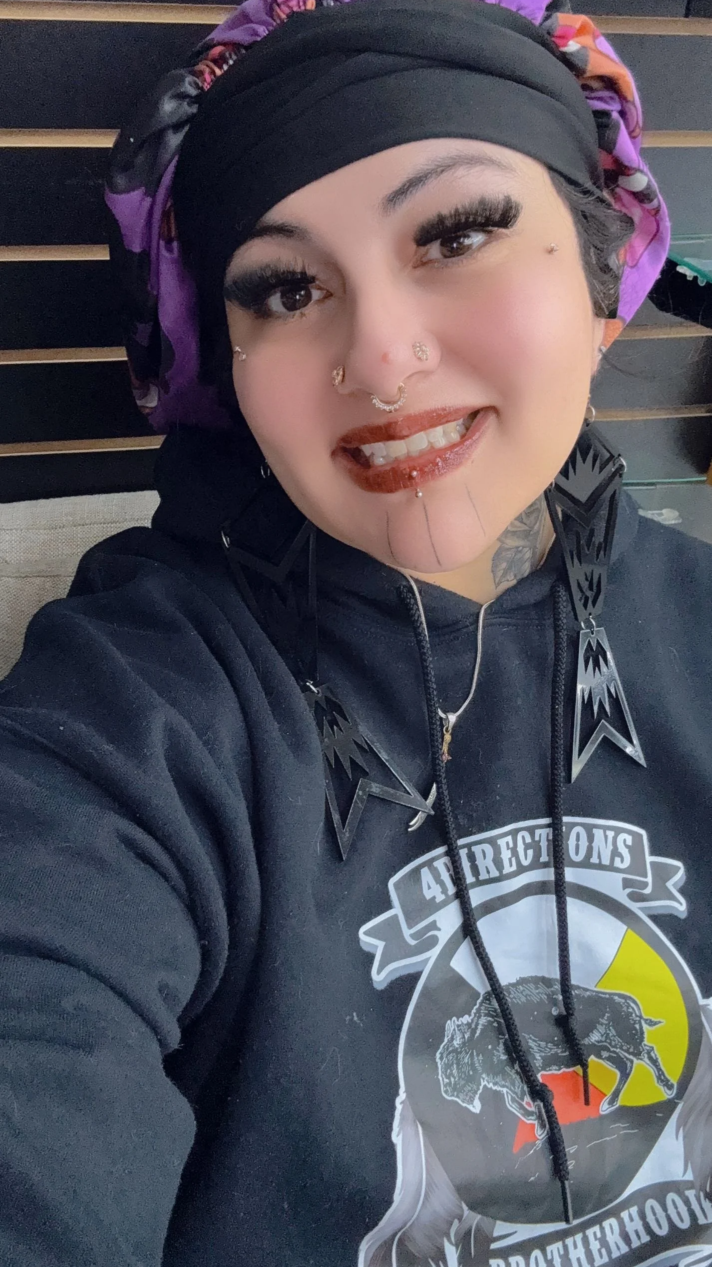 A woman smiling outdoors, wearing a black hijab with a colorful beaded flower clip on the side, makeup, and a plaid shirt with a black vest.