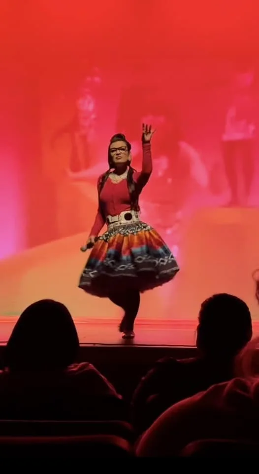 Performer on stage in colorful, patterned dress, raising hand in gesture, with red background and audience in foreground.