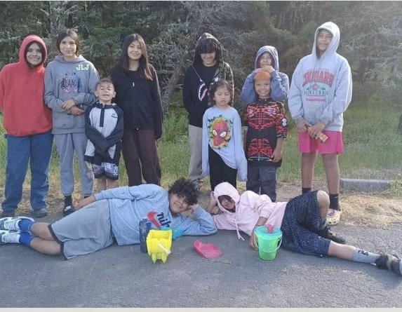 Group of children and teenagers posing outdoors on a paved surface, some lying down and others standing, with trees in the background.