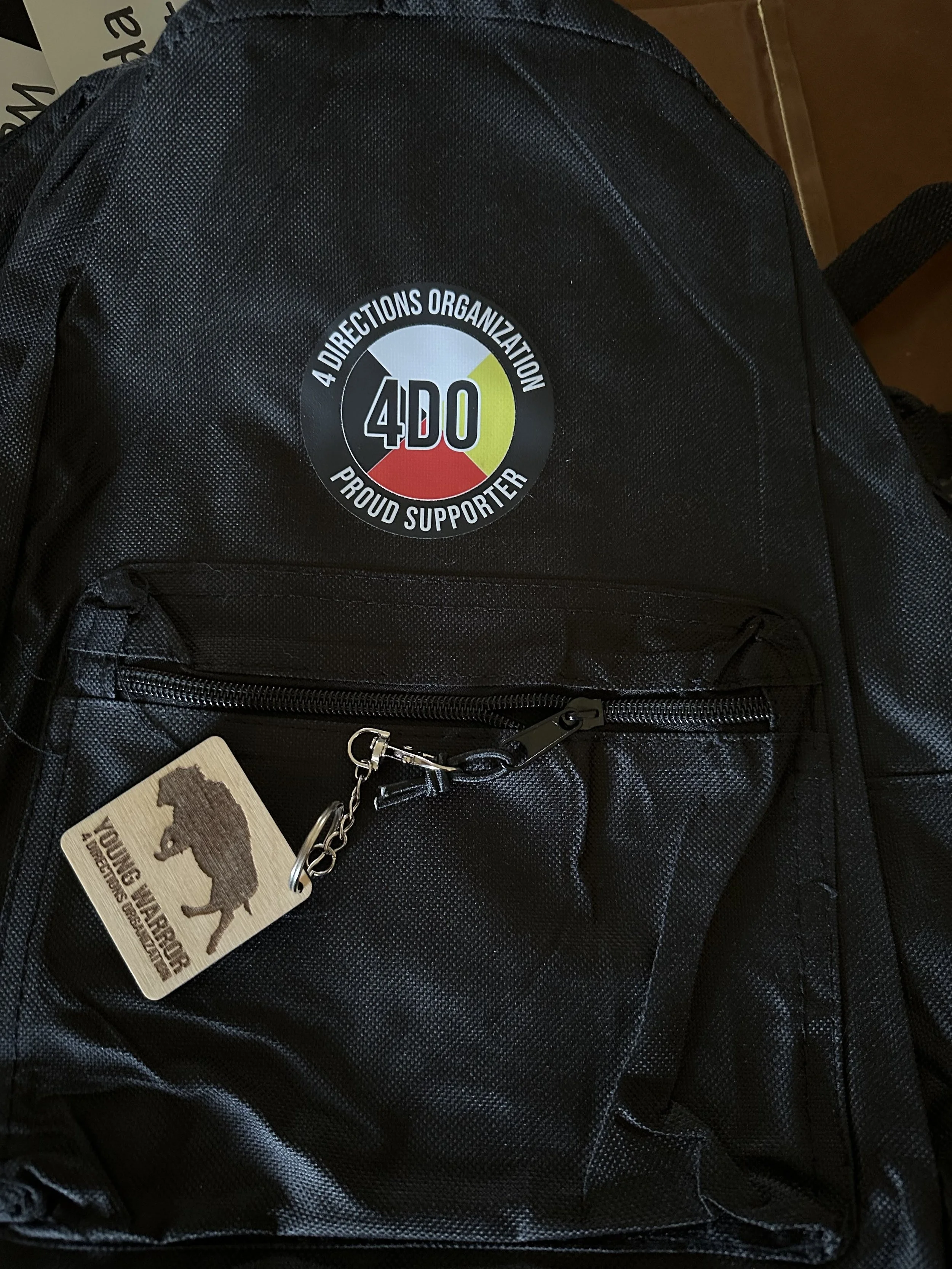 A black backpack with a round sticker that says '4 Directions Organization, Proud Supporter', featuring a color wheel with grey, green, yellow, and red sections. The backpack also has a keychain with a bear silhouette and a 'Young Bears' label.