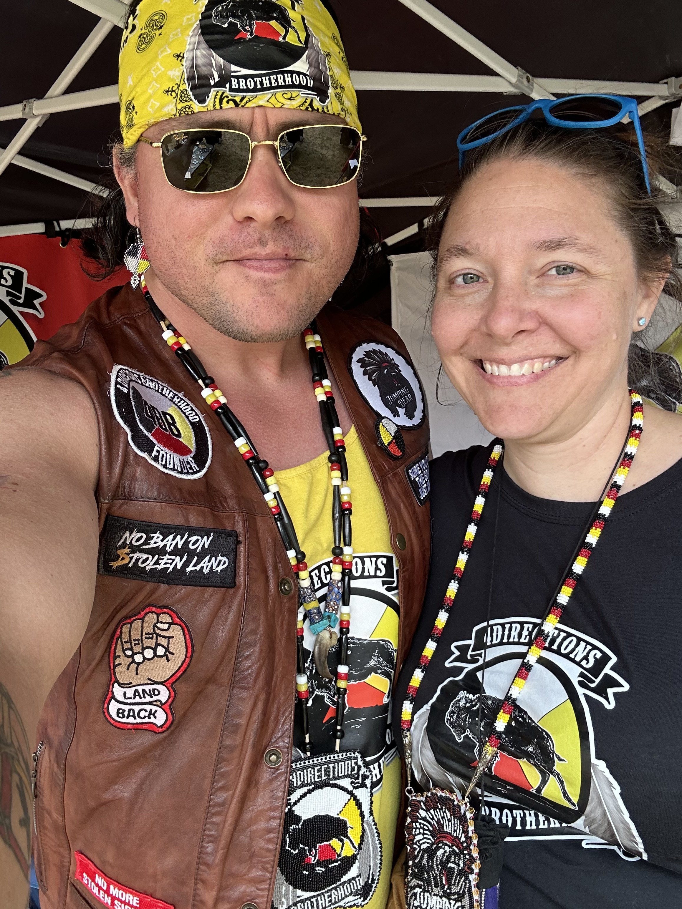 A man wearing sunglasses, a bandana with the Brotherhood logo, and multiple rings and bracelets, sitting with his hand resting on his chin, indoors in a cozy room with a lamp and window in the background.