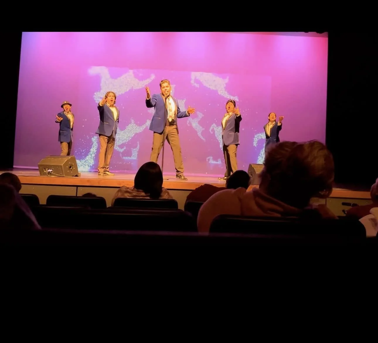 Stage performance with five performers dressed in blue blazers and khaki pants, standing in front of a pink backdrop with a dance-like design, audience seated in front.