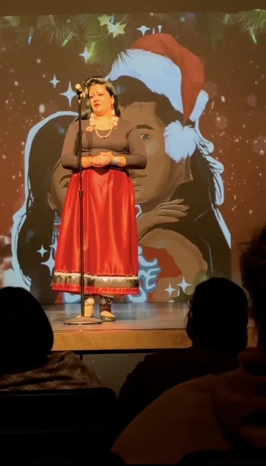 Woman wearing a black top and red skirt, standing on stage with a microphone. Behind her is a large digital backdrop of a girl wearing a Santa hat, surrounded by holiday decorations and stars.