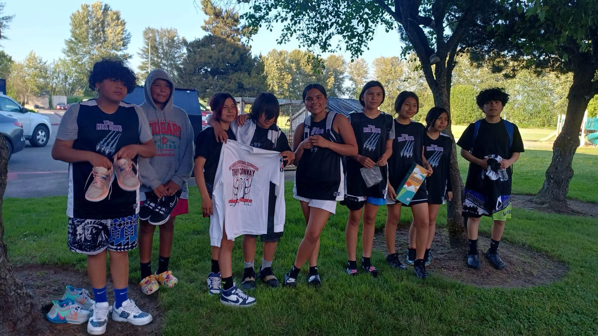 Group of young girls and boys in basketball uniforms standing outdoors on grass, some holding shoes and a basketball jersey, with trees and parked cars in the background.