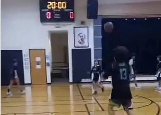 Indoor basketball court with a scoreboard showing 20:00 and tied score at 0-0, players on the court, and a man holding a basketball preparing to shoot.