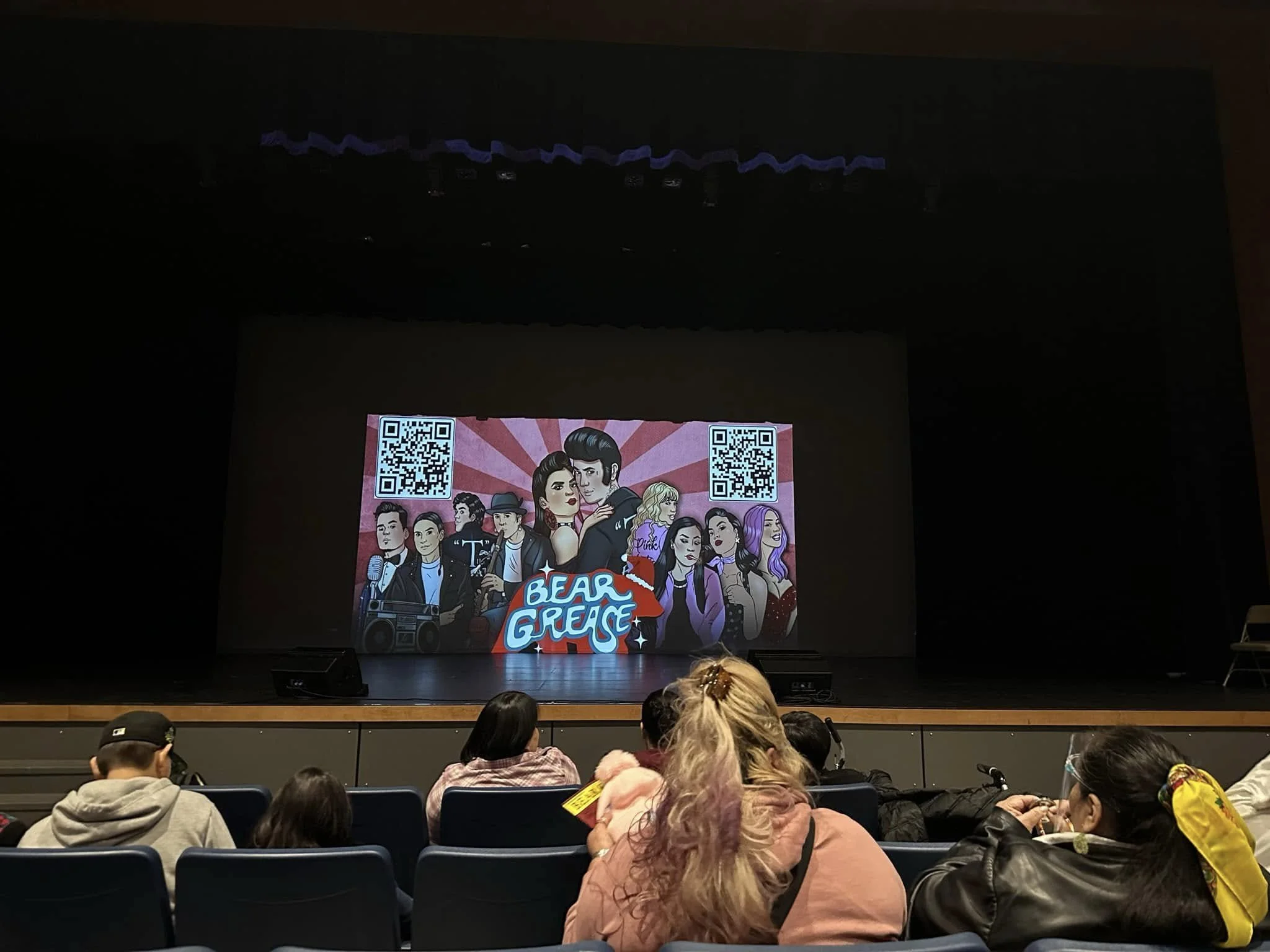 The image shows an auditorium with people seated and looking at a stage. On the stage, there is a large screen displaying an animated poster for 'Bear Grace' with illustrations of multiple characters and two QR codes.