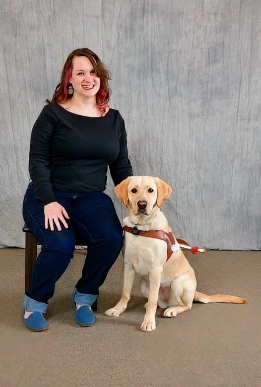 Leia is    smiling warmly and  sitting next to Havila, a yellow lab who is wearing a guide Dogs for the Blind harness
