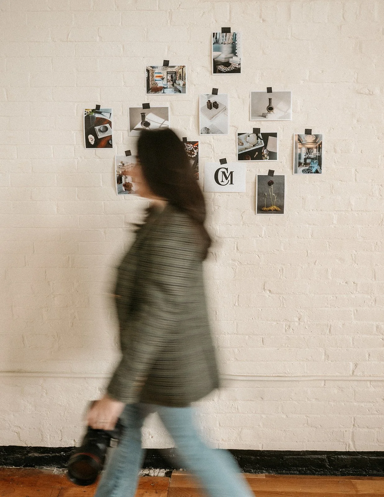 A person walking past a white brick wall with a photo collage taped to it, holding a camera in their right hand.