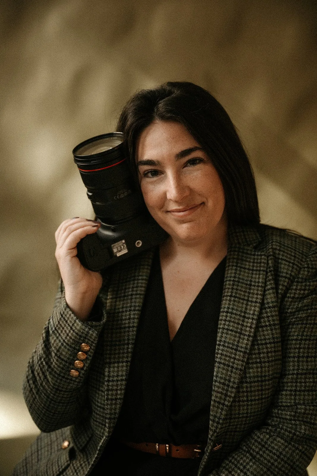 A woman with dark hair smiling and holding a professional camera lens to her ear as if it were a phone, wearing a checked blazer and black top.