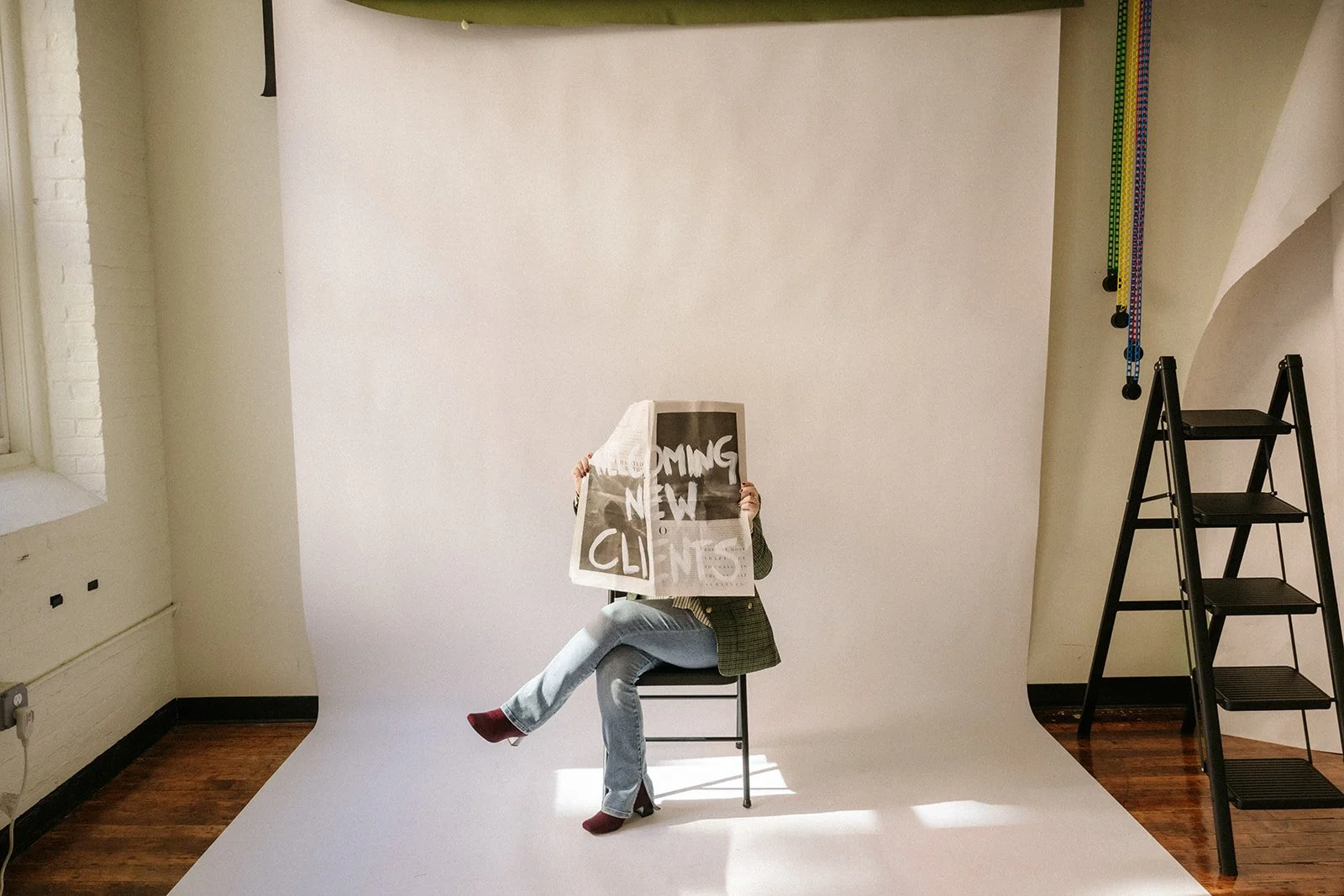 Person sitting on a chair holding a newspaper that covers their face, in a photography studio with a white backdrop and a step ladder.