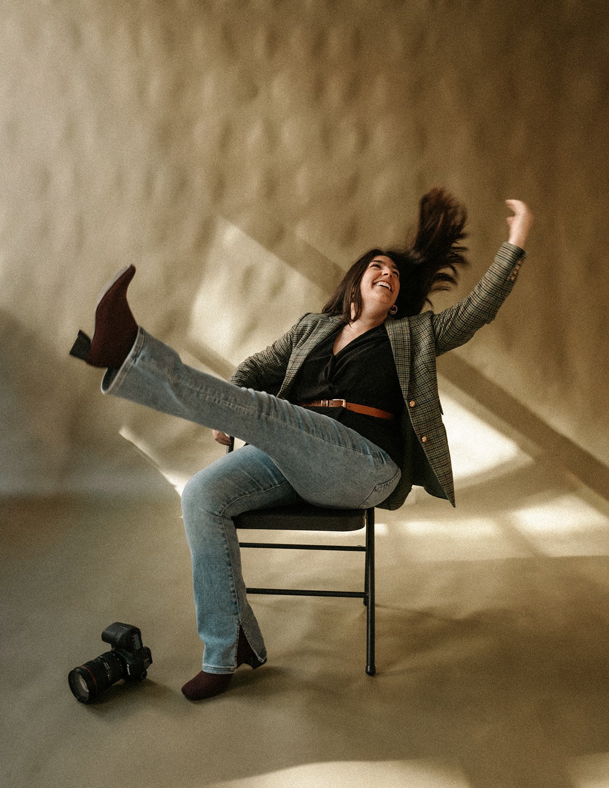 A woman sitting on a chair, laughing with her head thrown back, as her long hair swings in the air. She has one leg extended outward and wears a plaid blazer, black top, blue jeans, and maroon boots. A camera lies on the floor nearby.