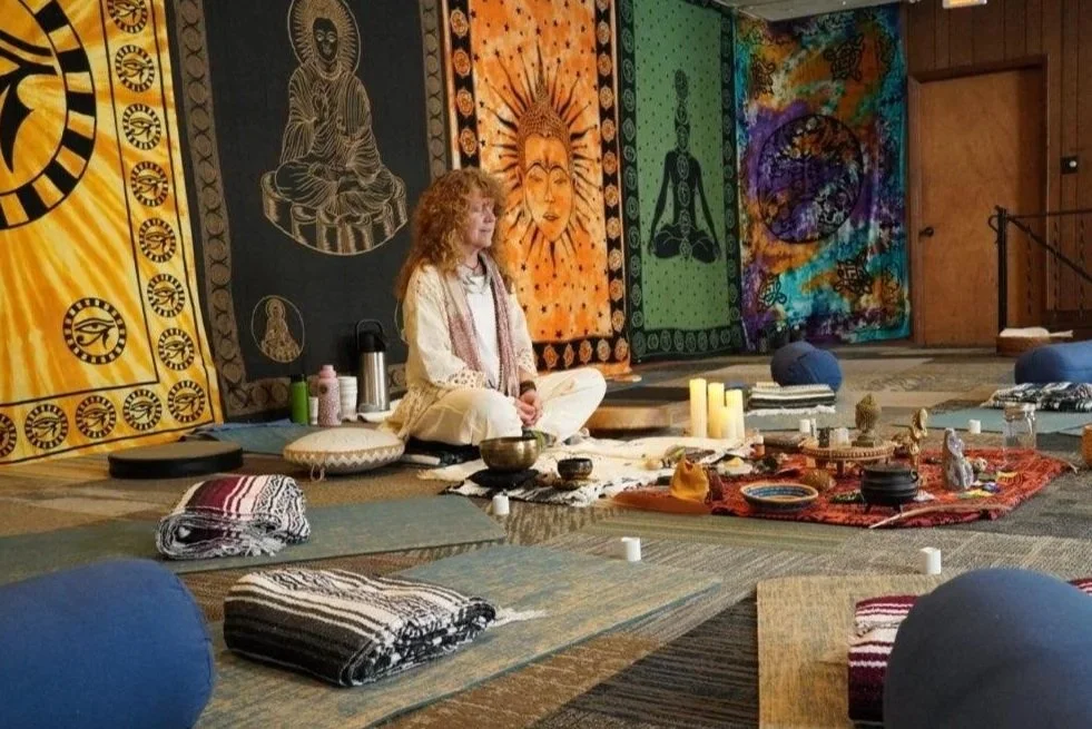 A woman with curly hair is sitting in a meditative pose on a mat in a room decorated with colorful tapestries featuring spiritual symbols and faces. There are candles, bowls, and other objects on rugs and mats around her, suggesting a spiritual or meditation session.