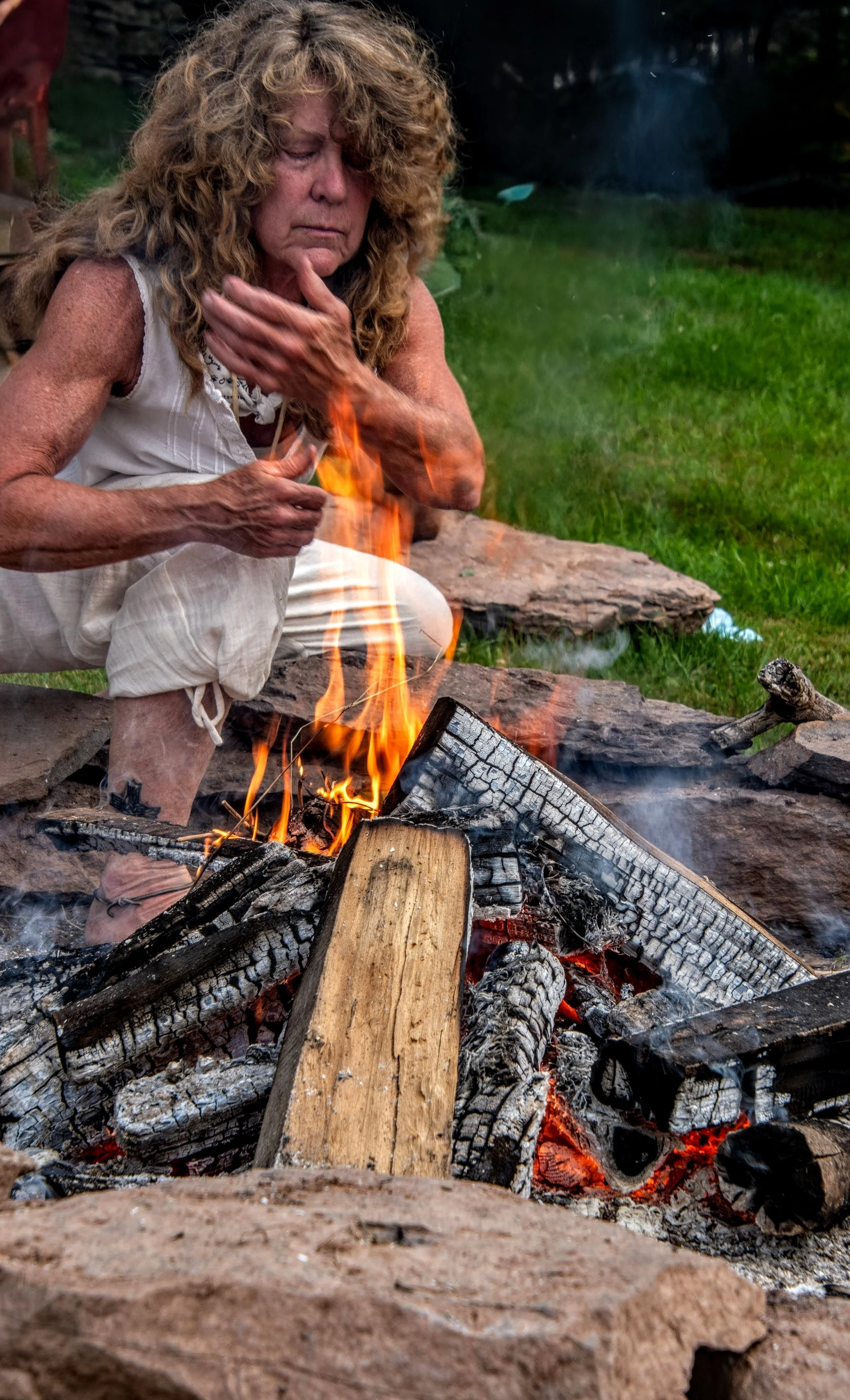 A woman with curly hair sitting by a campfire surrounded by rocks, holding her chin with her hand. The fire has burning logs and orange flames. A grassy area is visible in the background.