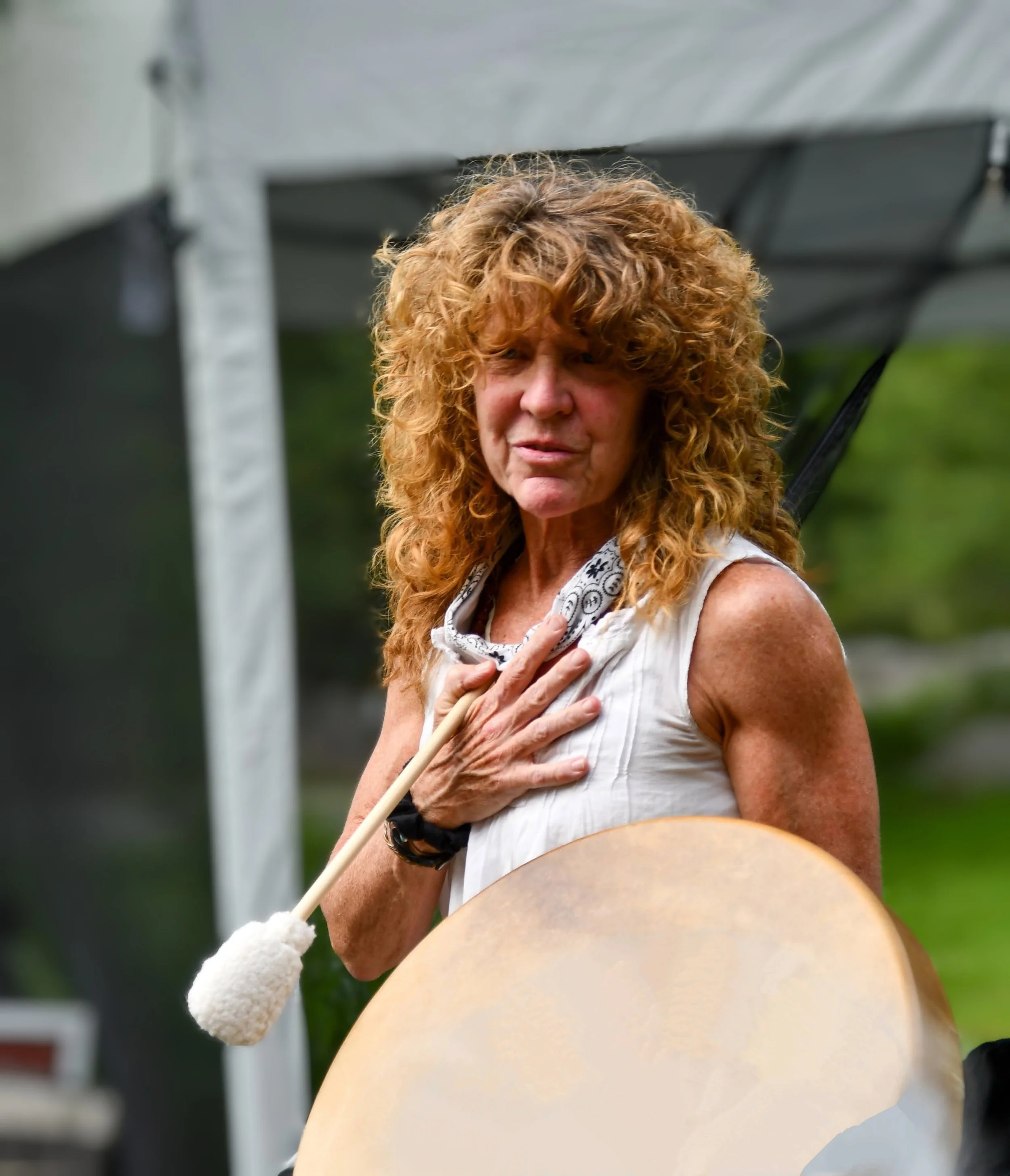 A woman with curly red hair holding a large gong, with her hand on her chest, outdoors with a tent and greenery in the background.