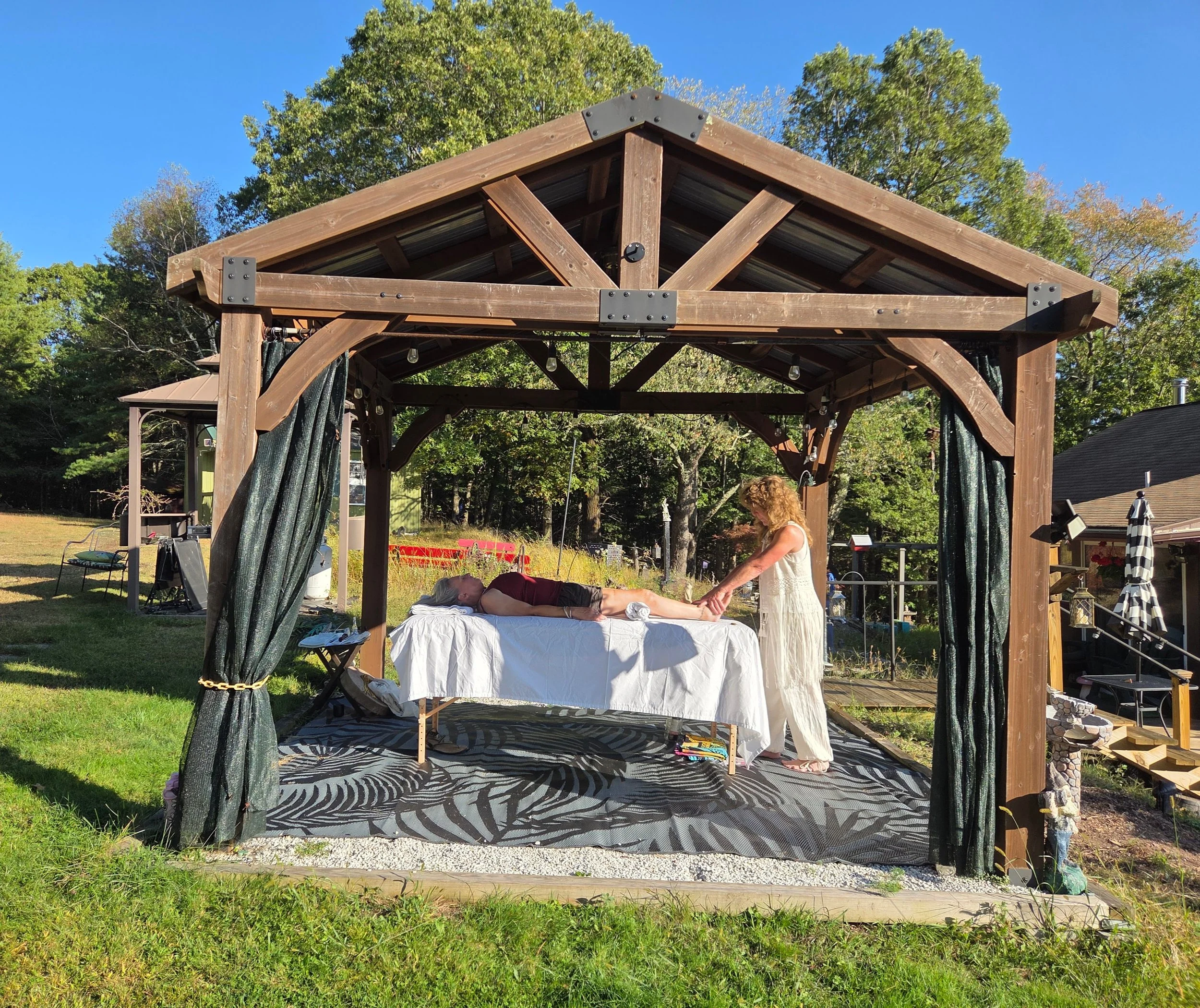 Outdoor massage setup under a wooden pergola with a therapist giving a massage to a person lying on a massage table, surrounded by trees and a grassy area.