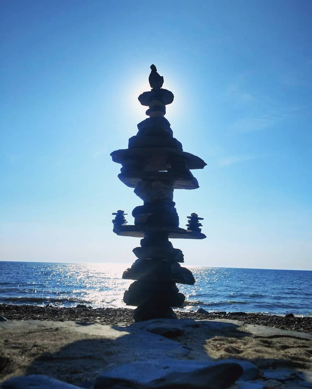 A stack of balanced rocks on a beach with the ocean in the background, and the sun positioned behind the top of the rock formation.