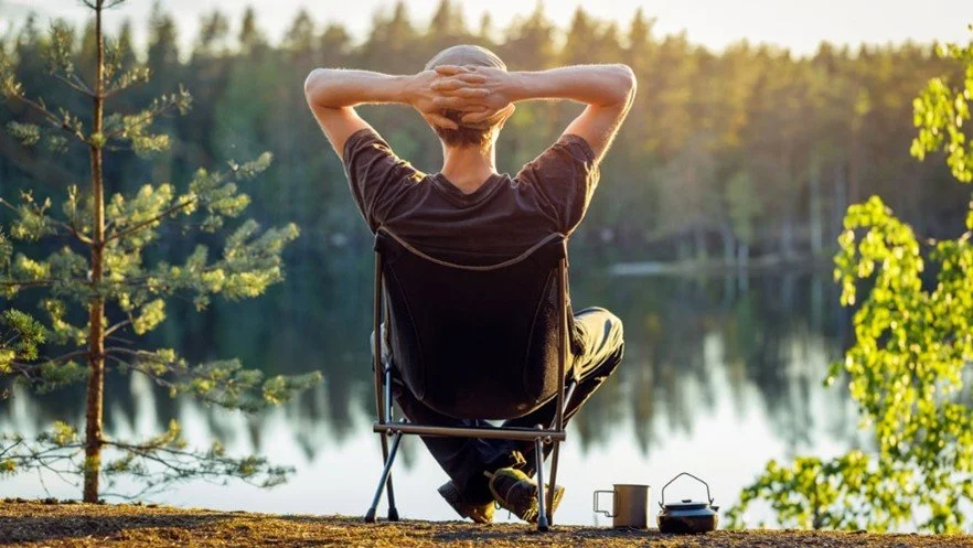 Person sitting on a portable chair near a lake with greenery, relaxing with hands behind head, during sunset or sunrise.