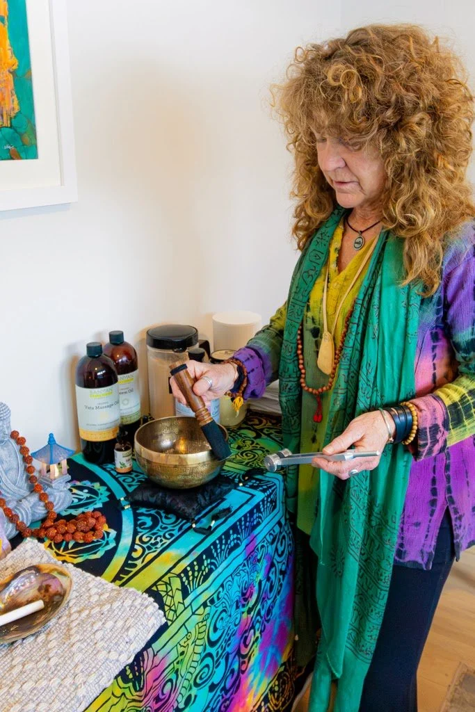 Woman with curly red hair wearing colorful clothing and jewelry, holding a small metal bowl and a wand, standing beside a table with spiritual and healing items, including bottles, beads, and a decorative cloth.