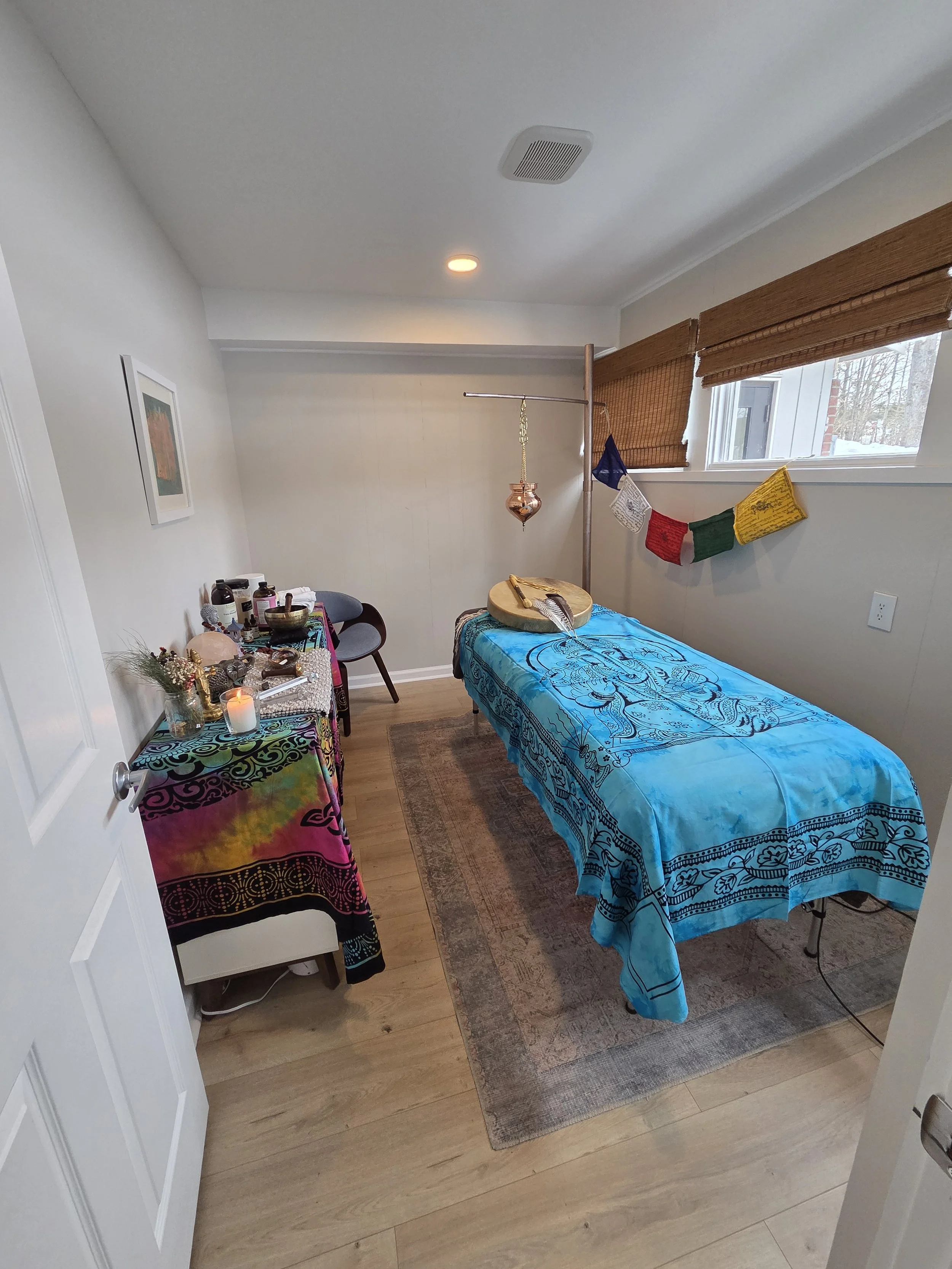 A massage room with a massage table covered in a blue cloth, a small table with spiritual items, a chair, and a window with bamboo blinds.
