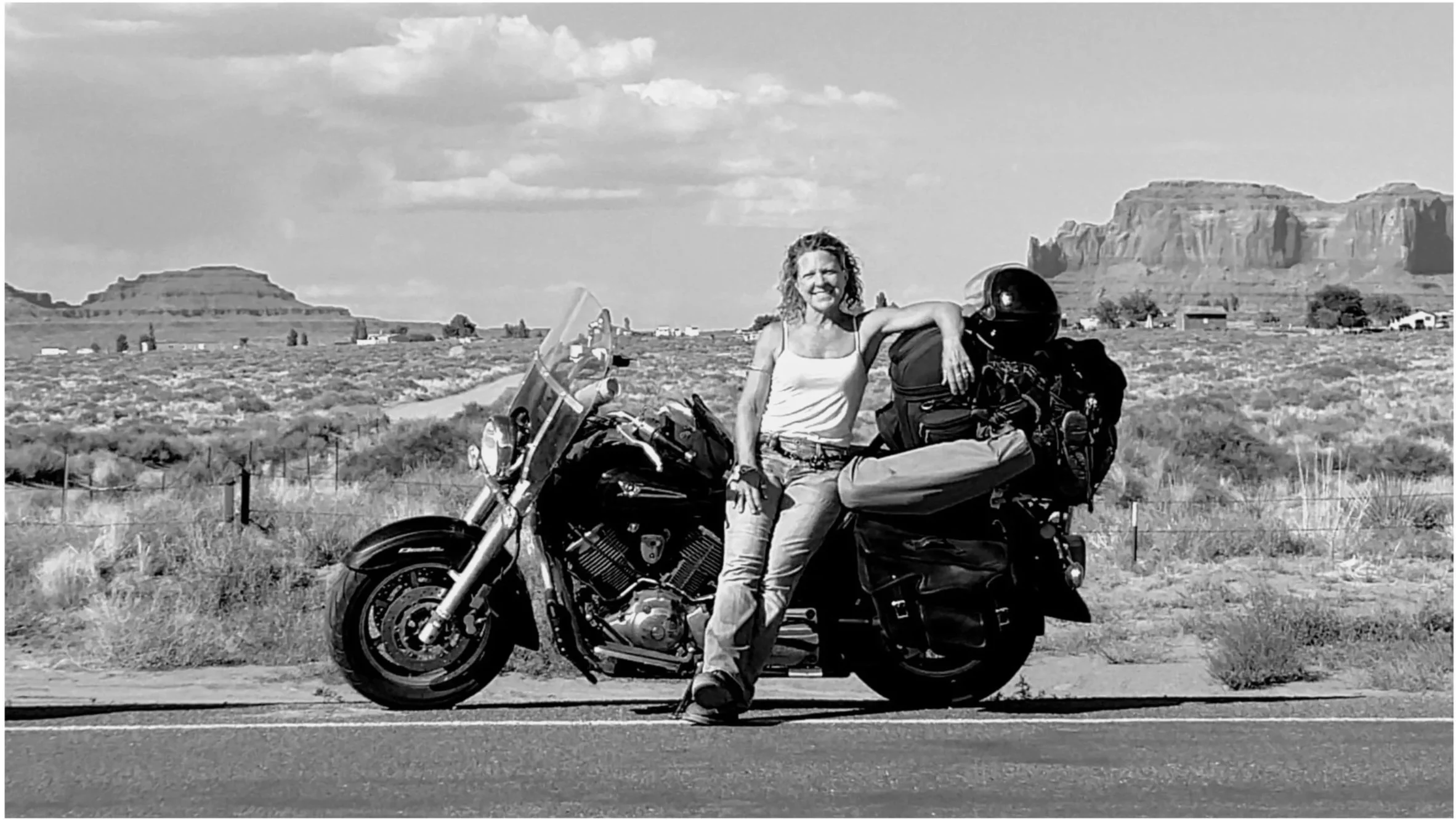 A woman with curly hair, wearing a tank top and jeans, sitting on a black motorcycle with luggage and a helmet, in a desert landscape with rock formations in the background.