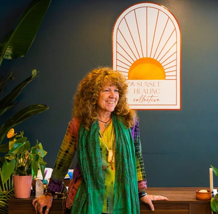 Woman with curly hair smiling, standing in front of a wall with a sunset sign, surrounded by potted plants.