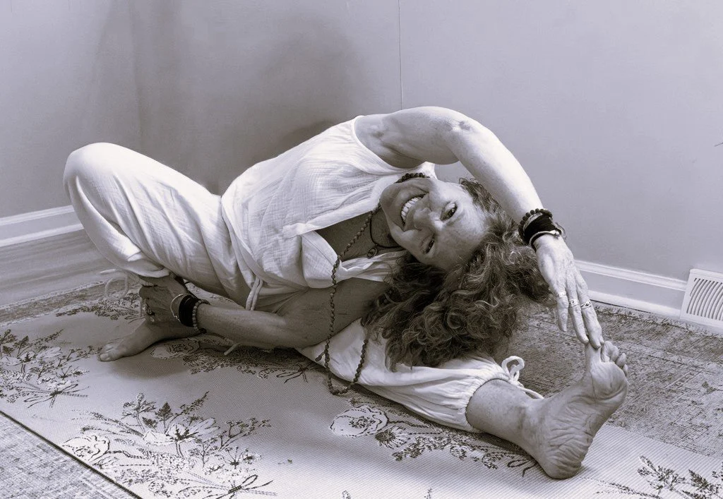 A woman with curly hair doing a seated yoga stretch on a rug, smiling, in a room with plain walls and a heating vent.