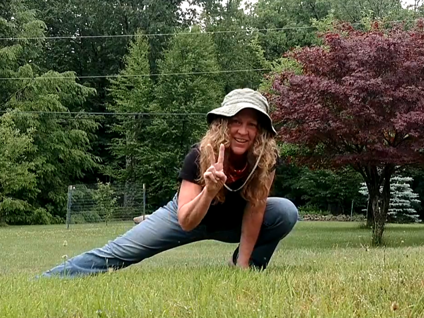 Woman in outdoor setting performing a split, wearing a hat, black shirt, and jeans, smiling and making a peace sign.