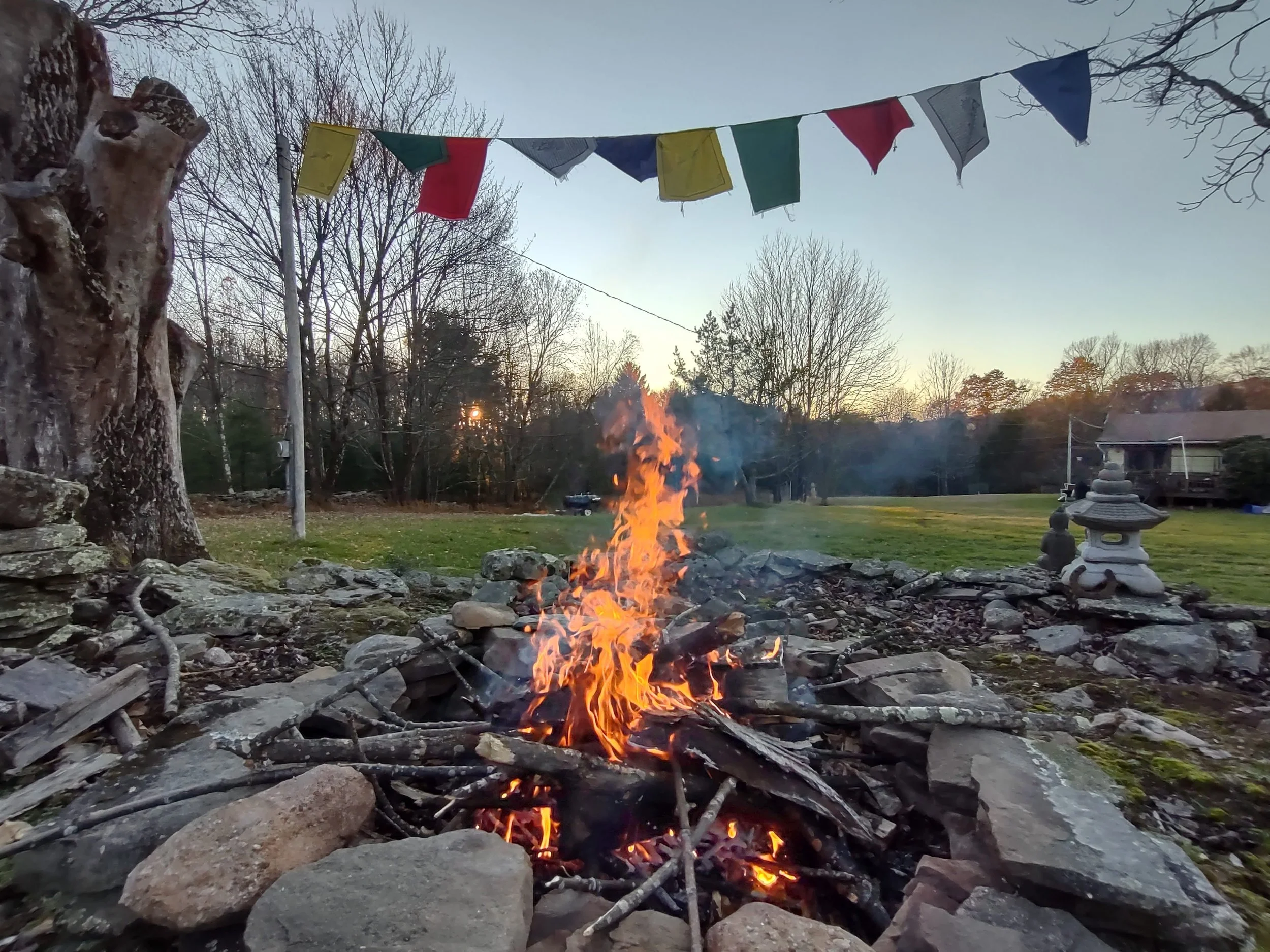 A backyard scene at sunset with a small campfire surrounded by rocks, a string of colorful prayer flags hanging overhead, trees in the background, and a decorative stone lantern on the right.