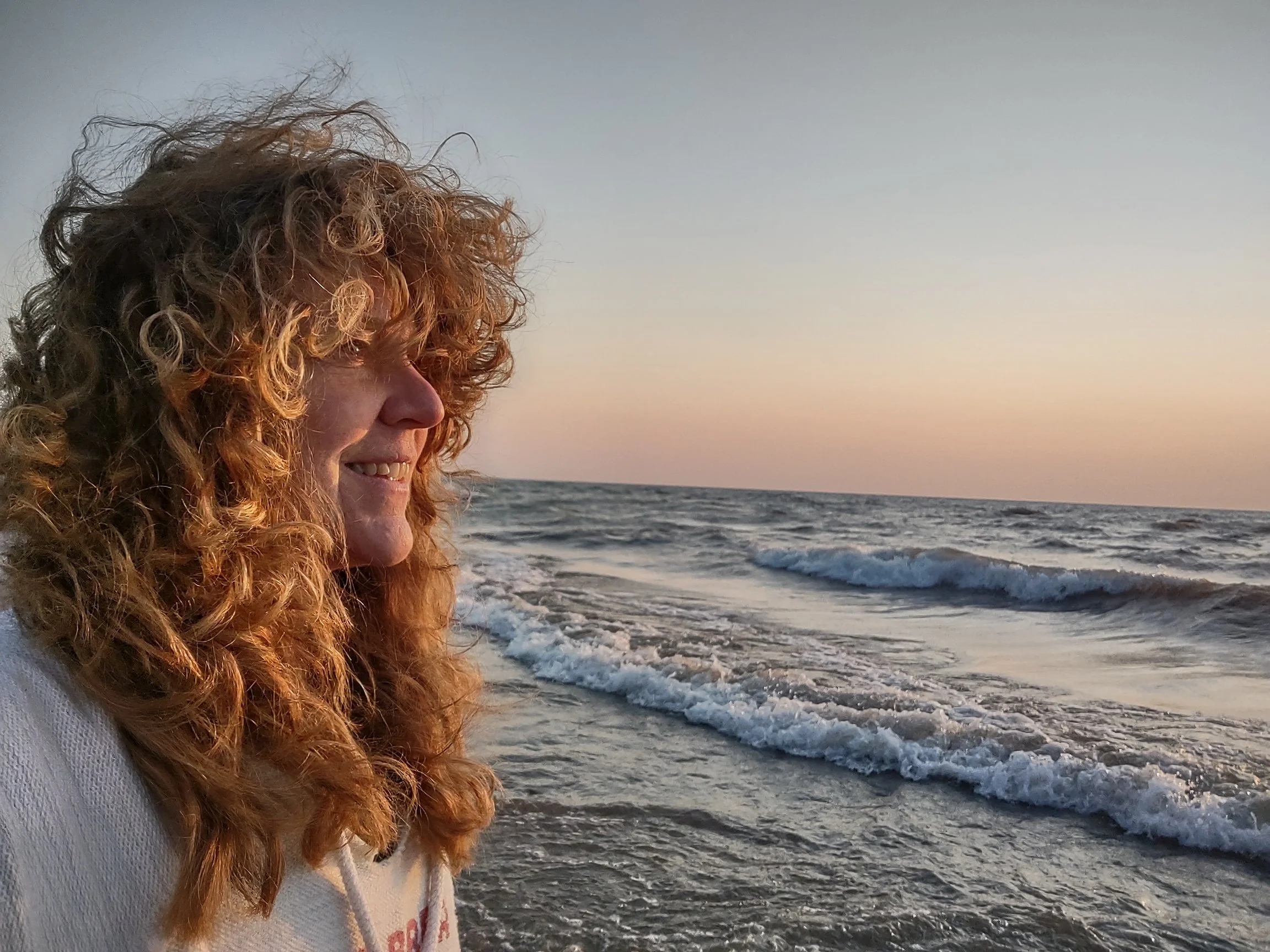 A woman with curly red hair smiling at the beach during sunset, standing near the shoreline with gentle waves in the background.