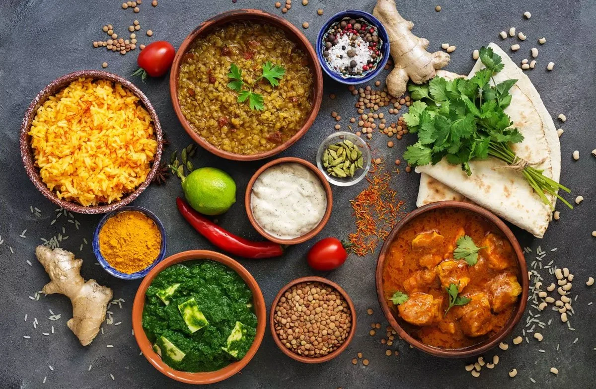 A variety of Indian dishes and spices arranged on a dark surface, including bowls of yellow rice, lentil curry, vegetable curry, mint chutney, yogurt sauce, turmeric powder, and fresh herbs, with ginger, chili, tomatoes, and spices scattered around.