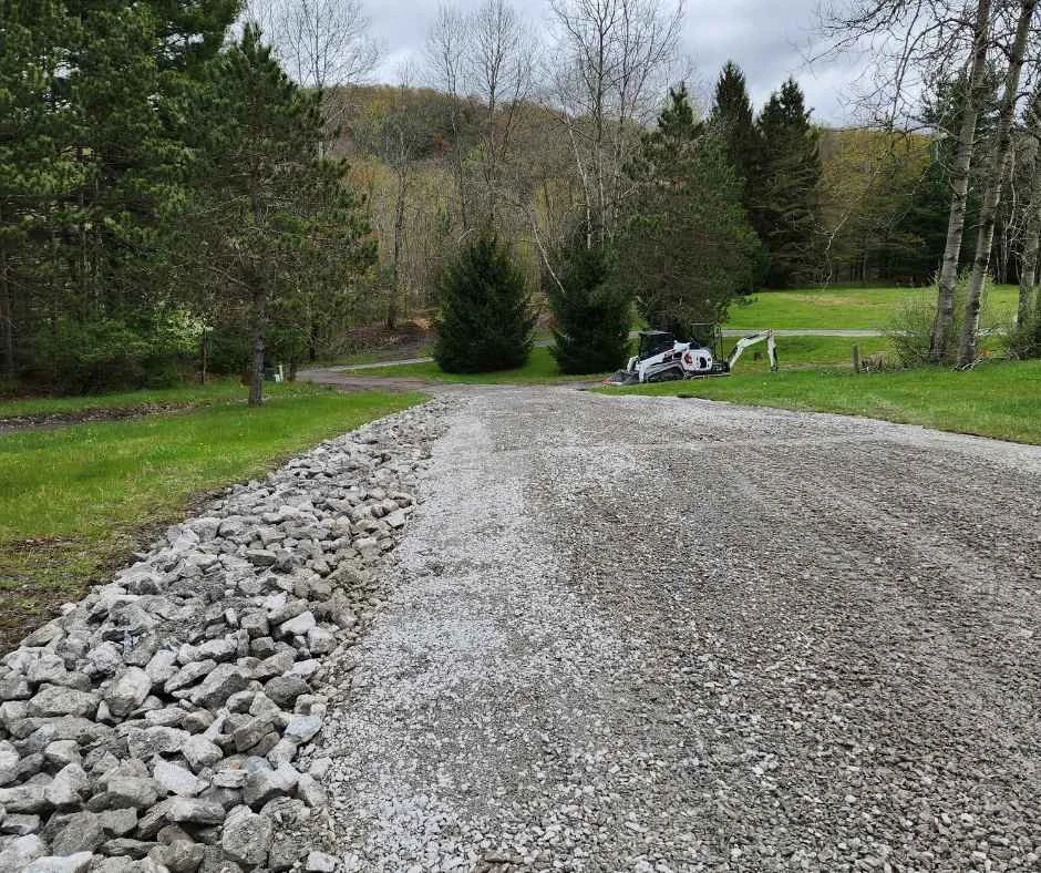A gravel driveway with rocks on the left side, leading to a grassy area with trees. A construction vehicle is parked in the background near a small shed.