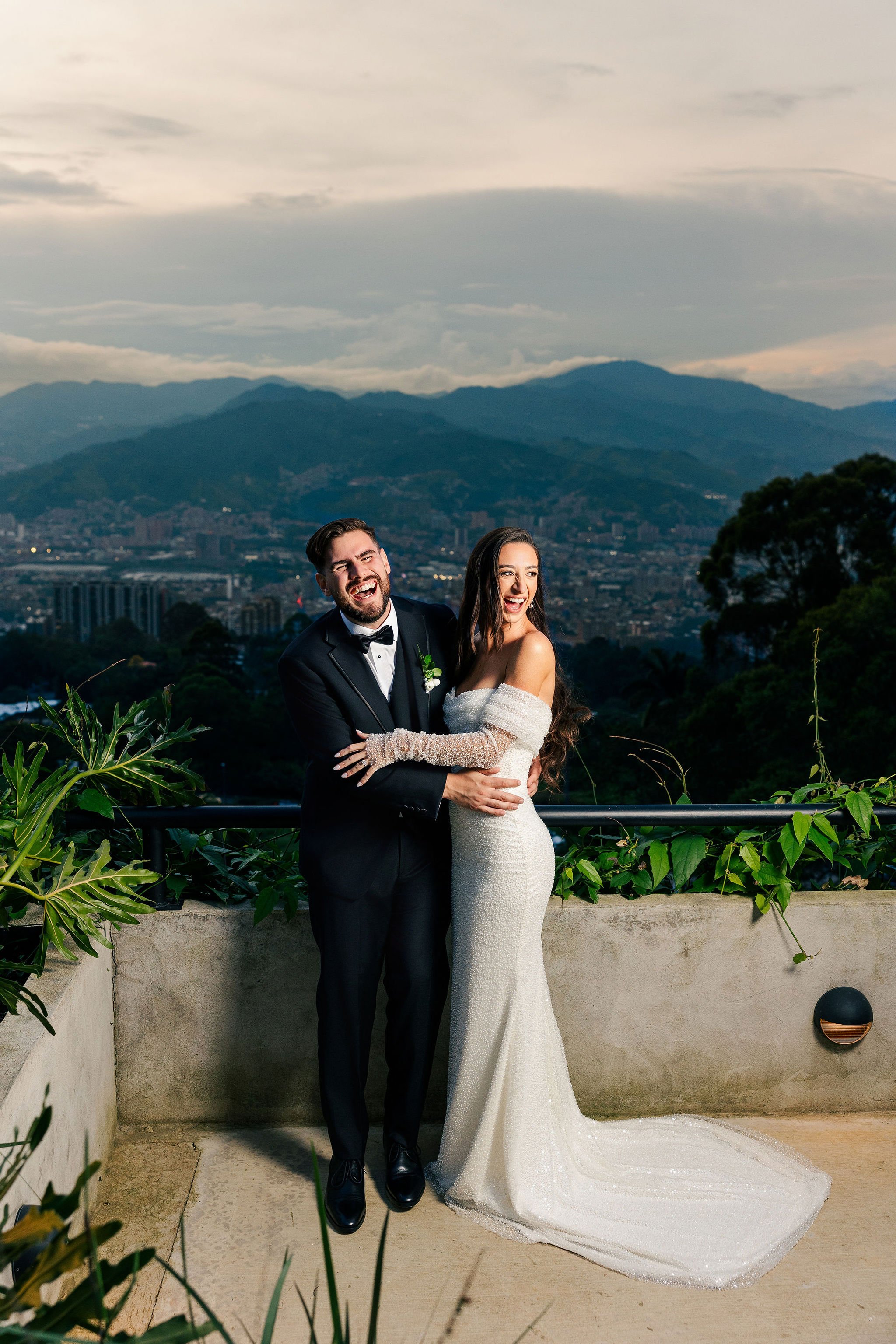 A newlywed couple dressed in wedding attire, standing on an outdoor terrace with a cityscape and mountain range in the background at sunset, smiling and laughing.