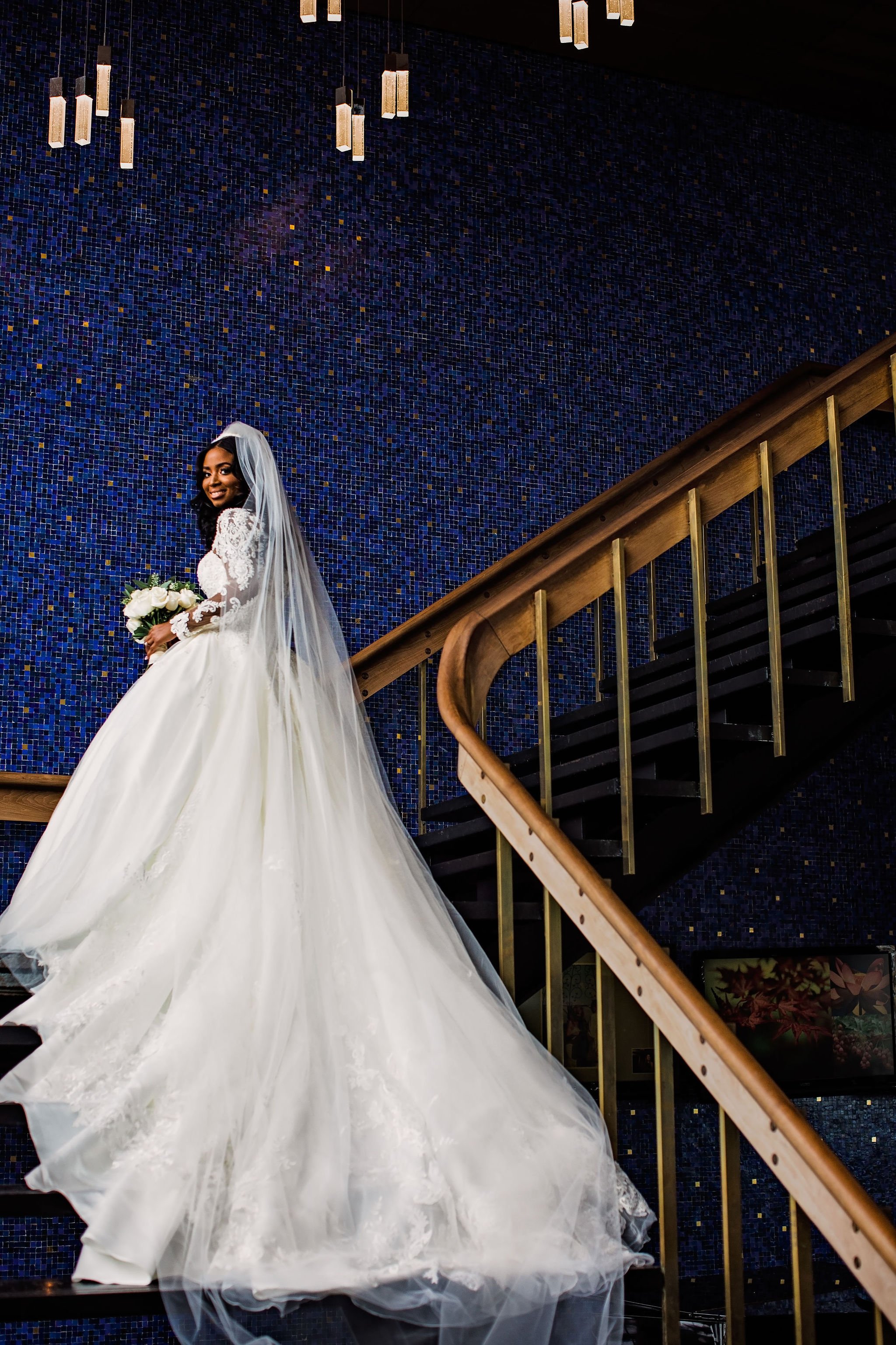 A bride in a white wedding gown and veil holding a bouquet of white roses standing on a staircase with a dark blue mosaic wall background.