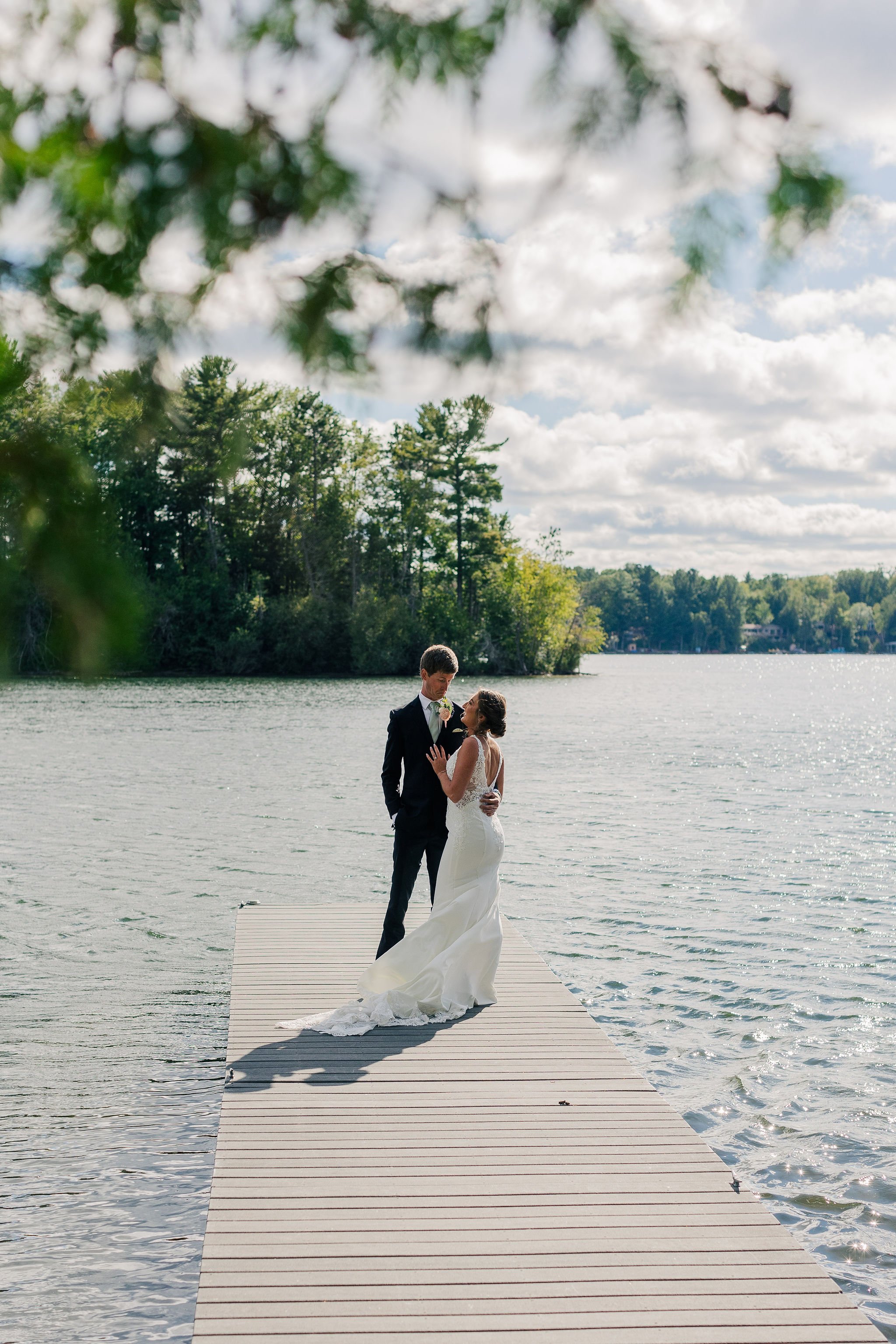 A bride and groom standing on a dock by a lake, embracing during their wedding day.