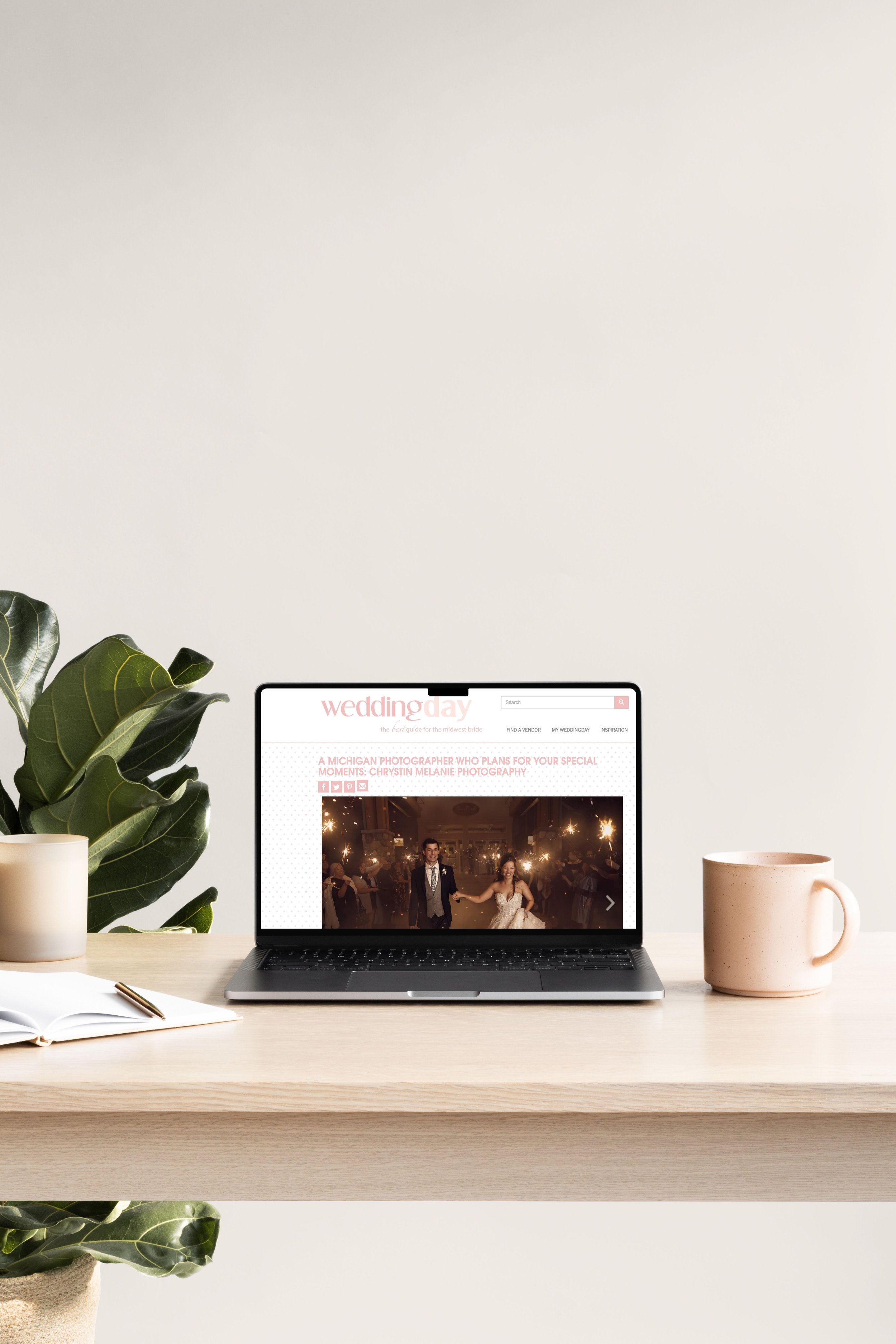 A wooden desk holding an open laptop displaying a wedding photography website, a potted plant, a closed notebook with a pen, and two cups, one cream-colored and one pink, set against a plain white wall.