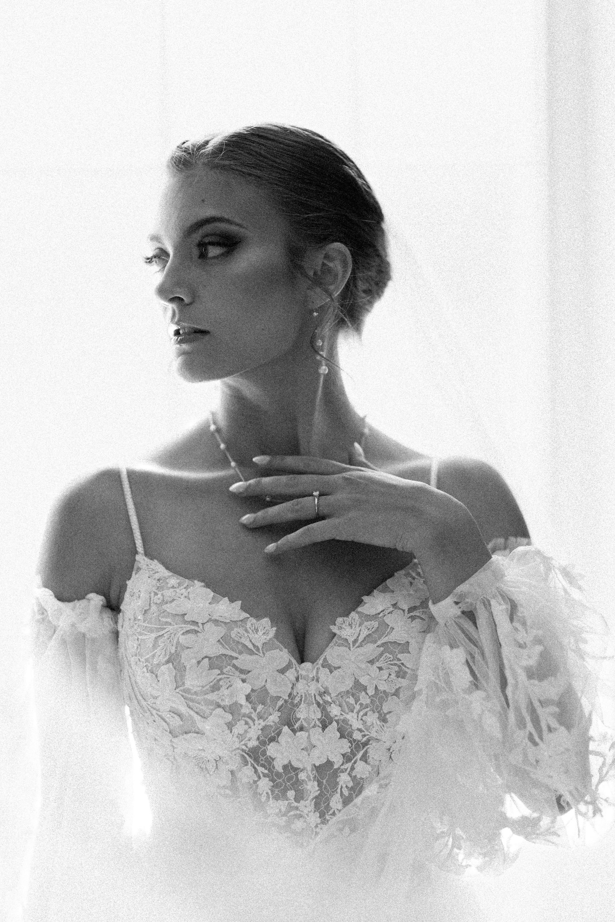 Black and white photo of a woman with styled hair, wearing a lace floral dress with off-the-shoulder sleeves, jewelry, and rings, looking to the side with her hand near her neck.